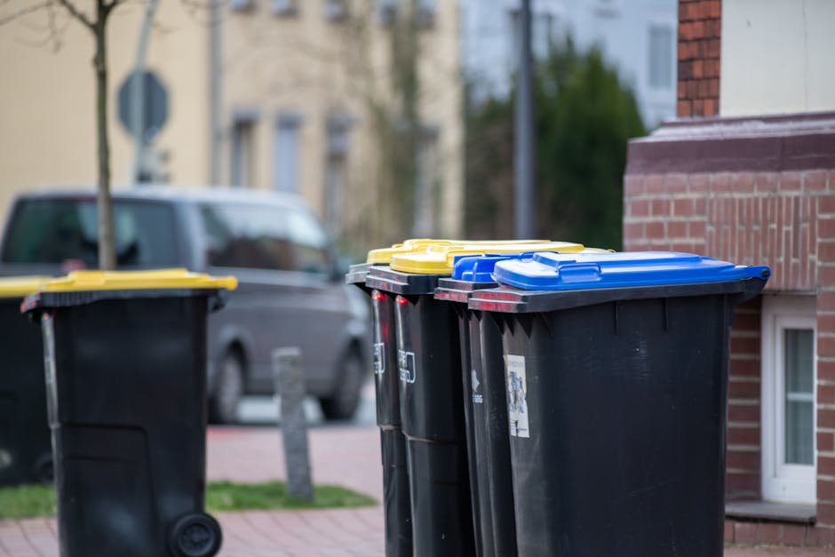 A row of large, upright wheelie bins lined up outdoors against a brick wall, with green bins at the front and blue and red bins positioned behind them. The green bins have textured, ribbed lids, while