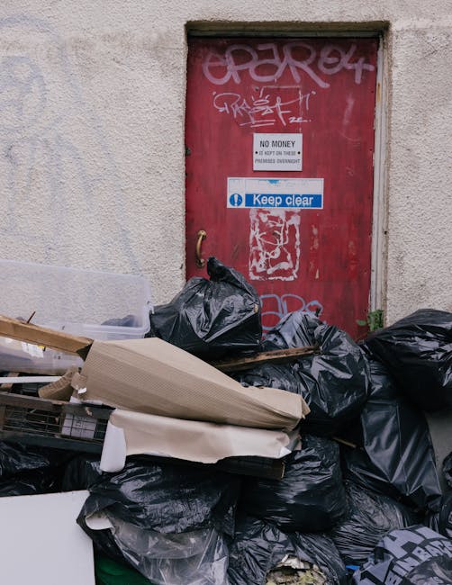 A worker wearing a yellow and red high-visibility jacket is operating a red rubbish collection truck on a roadside. The truck's rear compactor is open, revealing mechanical components and a metal hopp